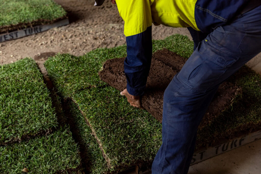 A person is shown from the waist down, bending over to lift and stack a square of fresh west turf.